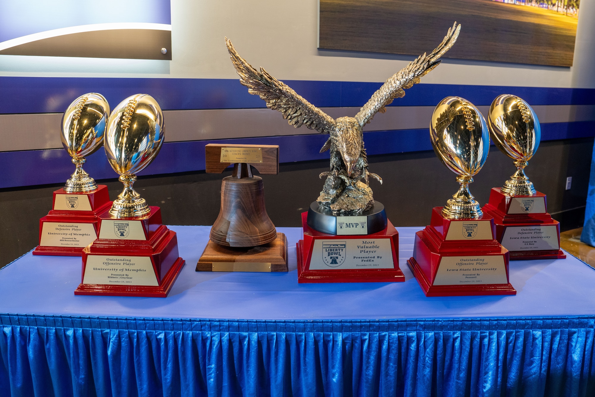 A close-up of the eagle trophy among other awards on a blue-clothed table. The trophies are labeled for various recognitions, such as MVP and Outstanding Player, with Liberty Bowl branding prominent in the background.