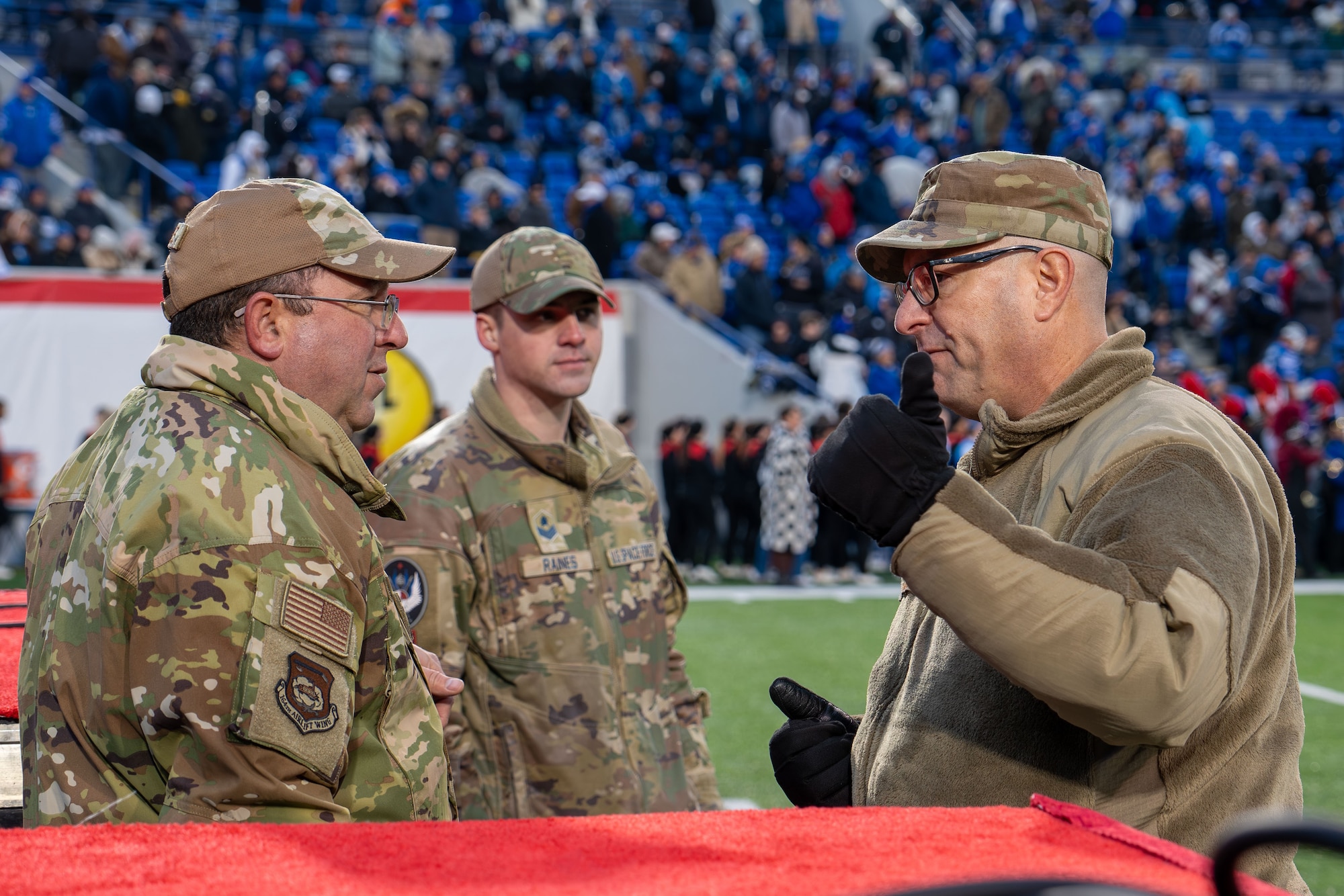 A photo of three military personnel in military uniform having a conversation on a football field. The man in the foreground is gesturing while speaking to the other. A third person is in the background, slightly out of focus. They are standing on a red carpet with a stadium full of spectators in the background.