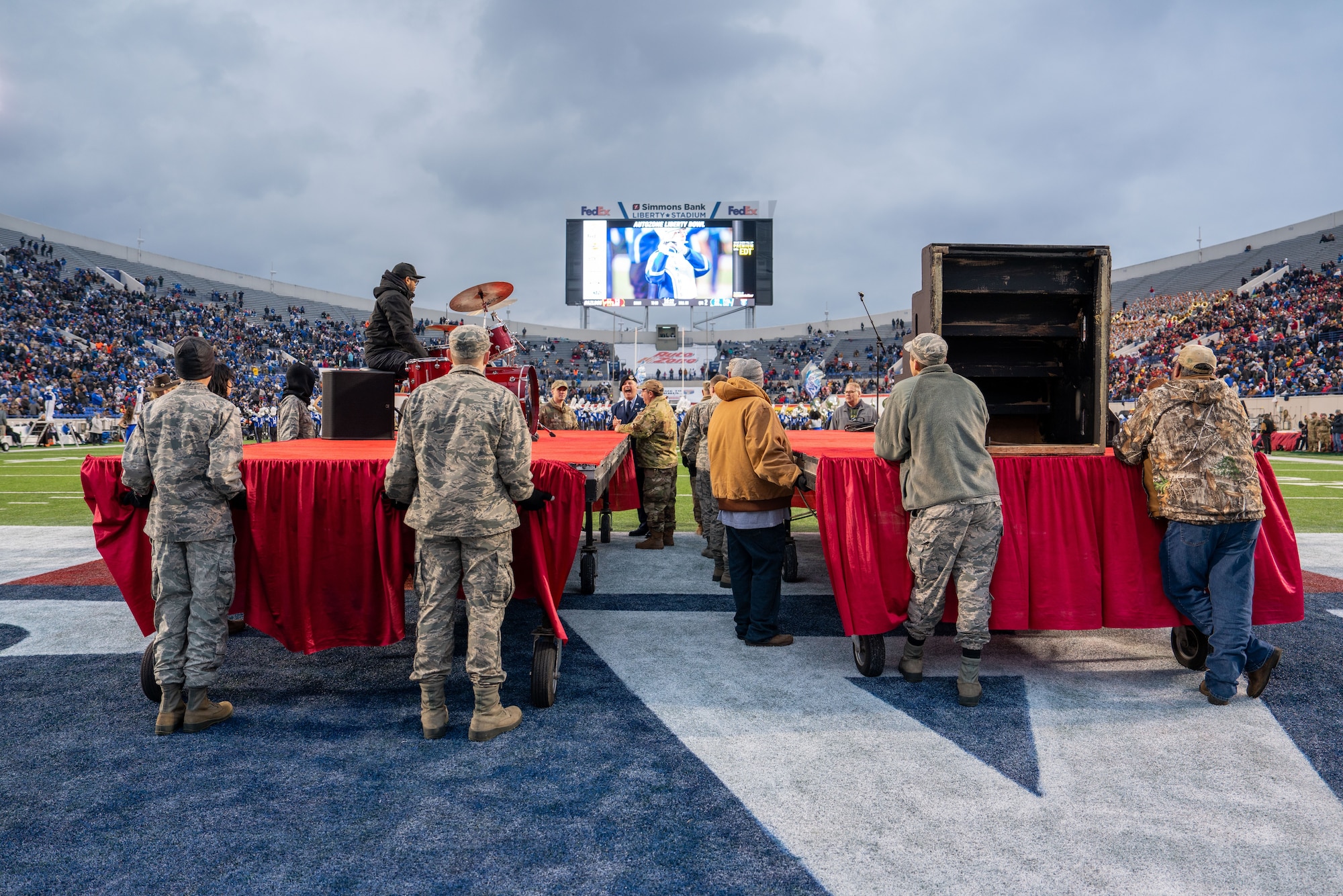 An image of a group of military personnel in uniform and civilians standing behind a red-covered stage on a football field. In the background, there is a band playing and a jumbotron showing close-ups. They were in preparation for the halftime performance, in which they had to rushe two stages onto the football field in time for a performance by the Memphis group, Bar-Kays.