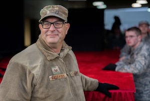 An image of a smiling man in military fleece gear with glasses and a cap stands by a red-draped stage, looking directly at the camera with a soft focus on the background where other military personnel can be seen.
