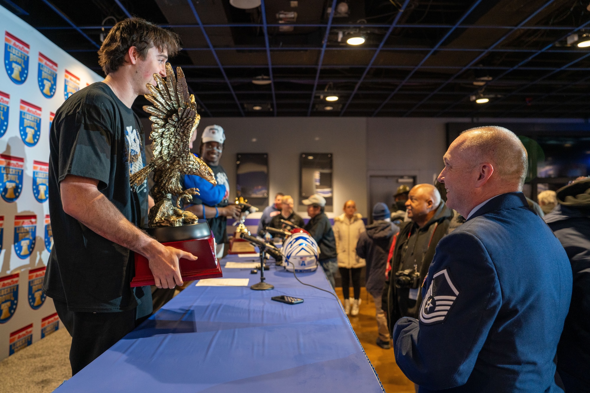 A photo of a man holding an eagle trophy, talking with a person in a U.S. Air Force dress uniform, while people around him are engaged in conversation or looking at the trophy. The setting suggests a casual, celebratory atmosphere with the Liberty Bowl signage in the background.
