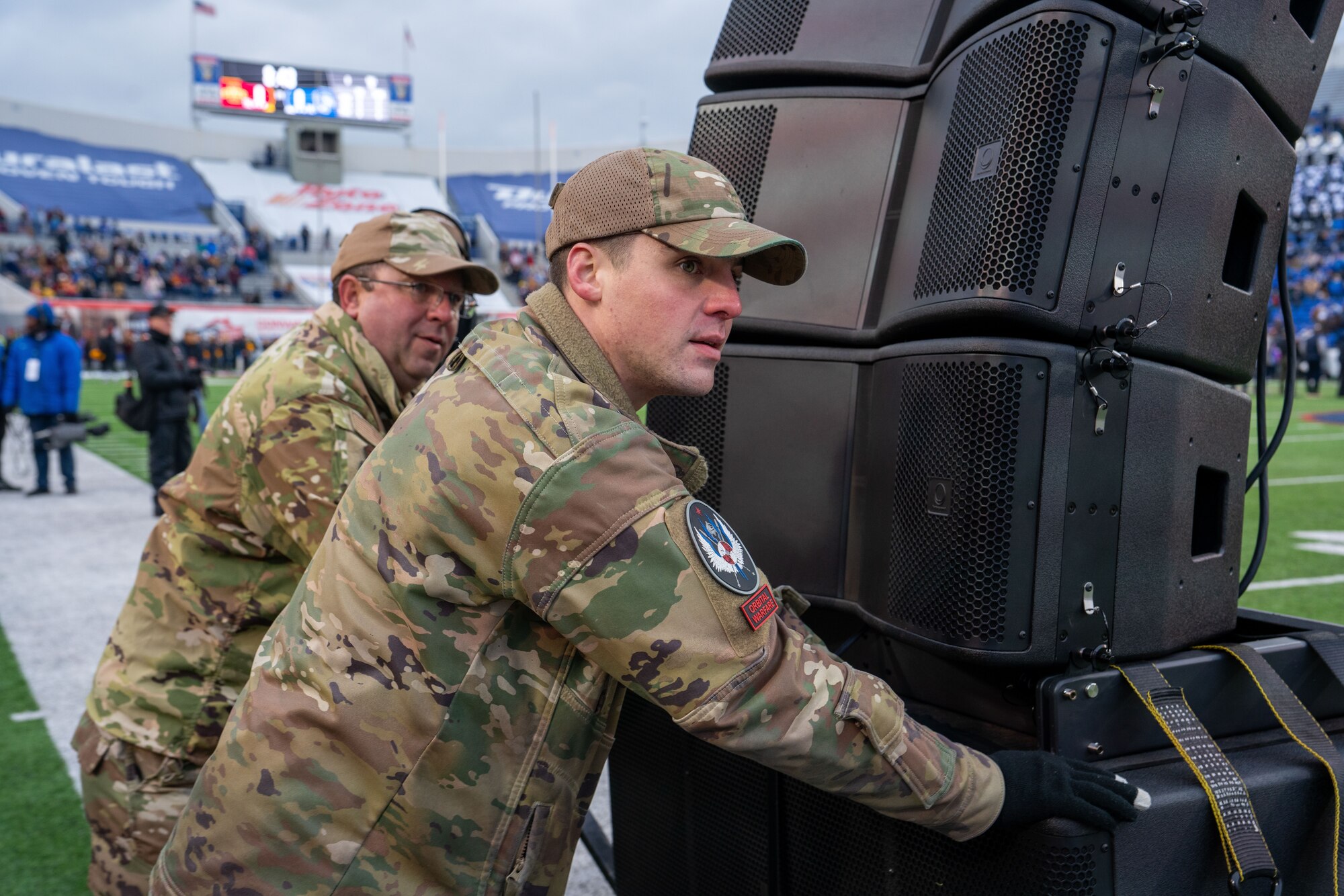 A photo of two military personnel in uniform who are pushing a large speaker on a football field. They are setting up audio equipment for an event, with a sports stadium in the background.