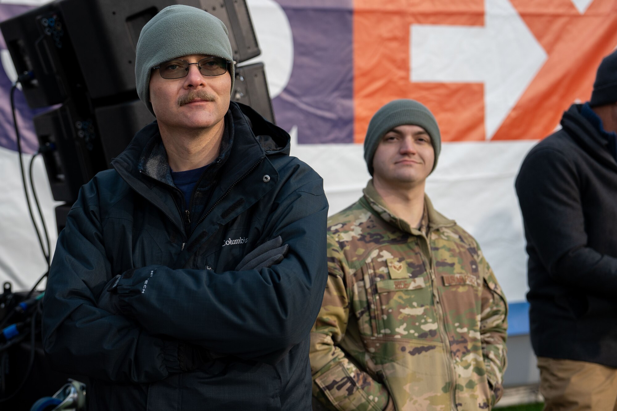An image of two individuals, one in a winter jacket, standing with arms crossed, and beanie and the other in a military jacket, both looking off to the side. They are near audio equipment for an event.