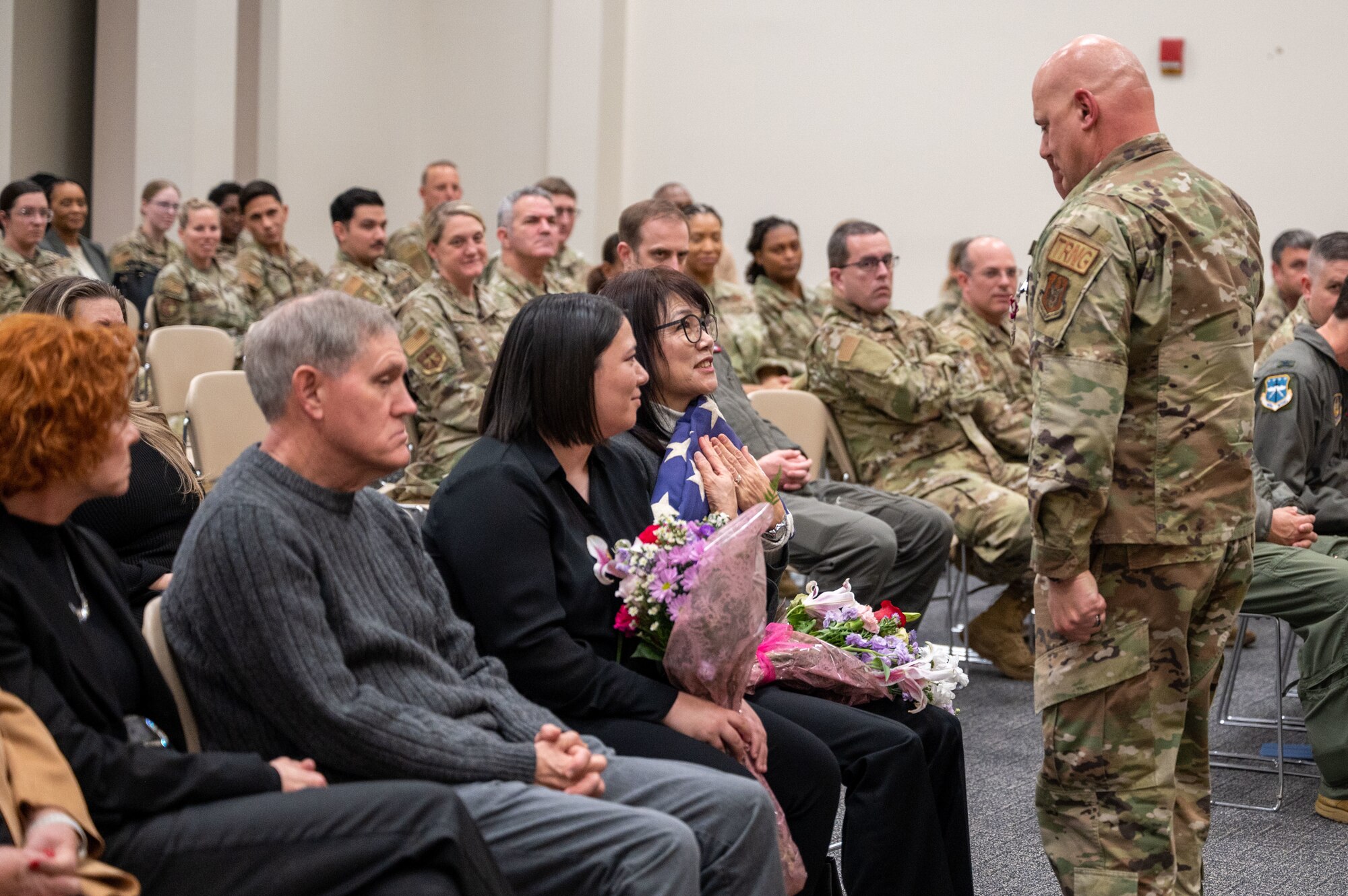 People sit, a woman holds flowers and a folded flag while Blanchard stands in front of her saluting it