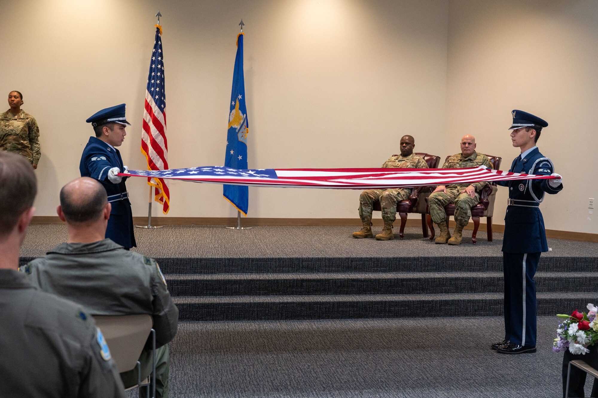 Two Honor Guard Airmen stretch an American Flag out between them. Blanchard and another member sit in the background watching