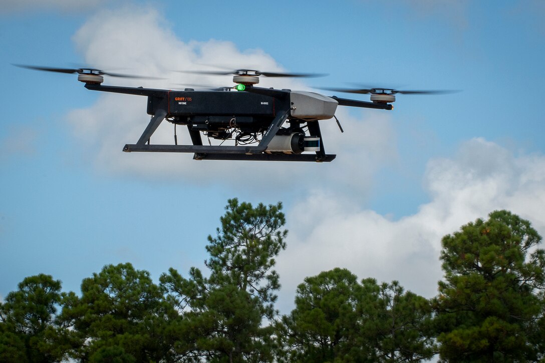 A drone flies in blue sky above a tree line in the distance.