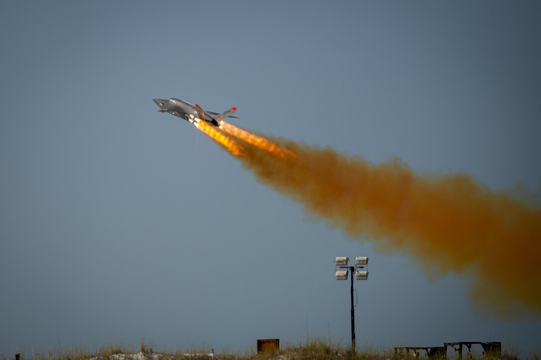 A XQ-58A Valkyrie launches into the sky, creating flames and a trail of orangish smoke.