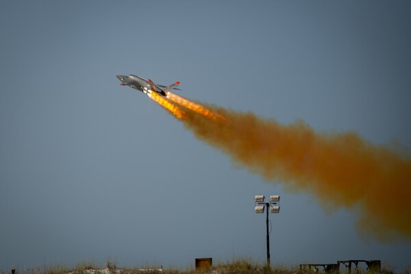 A XQ-58A Valkyrie launches into the sky, creating flames and a trail of orangish smoke.
