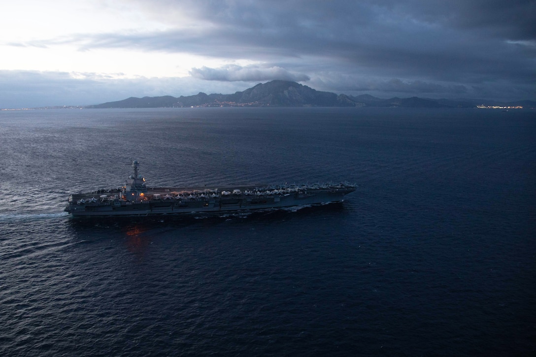 An aircraft carrier sails off a mountainous coast at twilight.