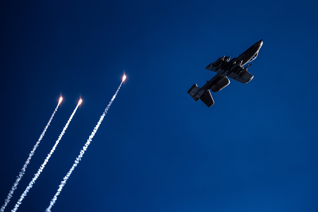 A military aircraft deploys three flares that can be seen against a deep, blue sky.