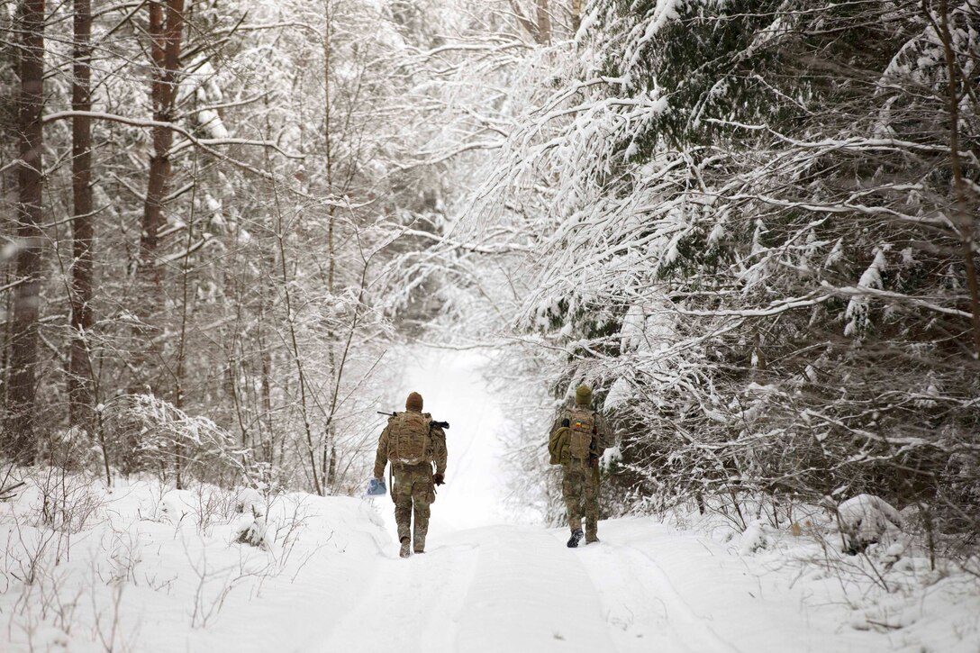 U.S. and Romanian soldiers wearing tactical gear walk down a snowy path surrounded by trees.