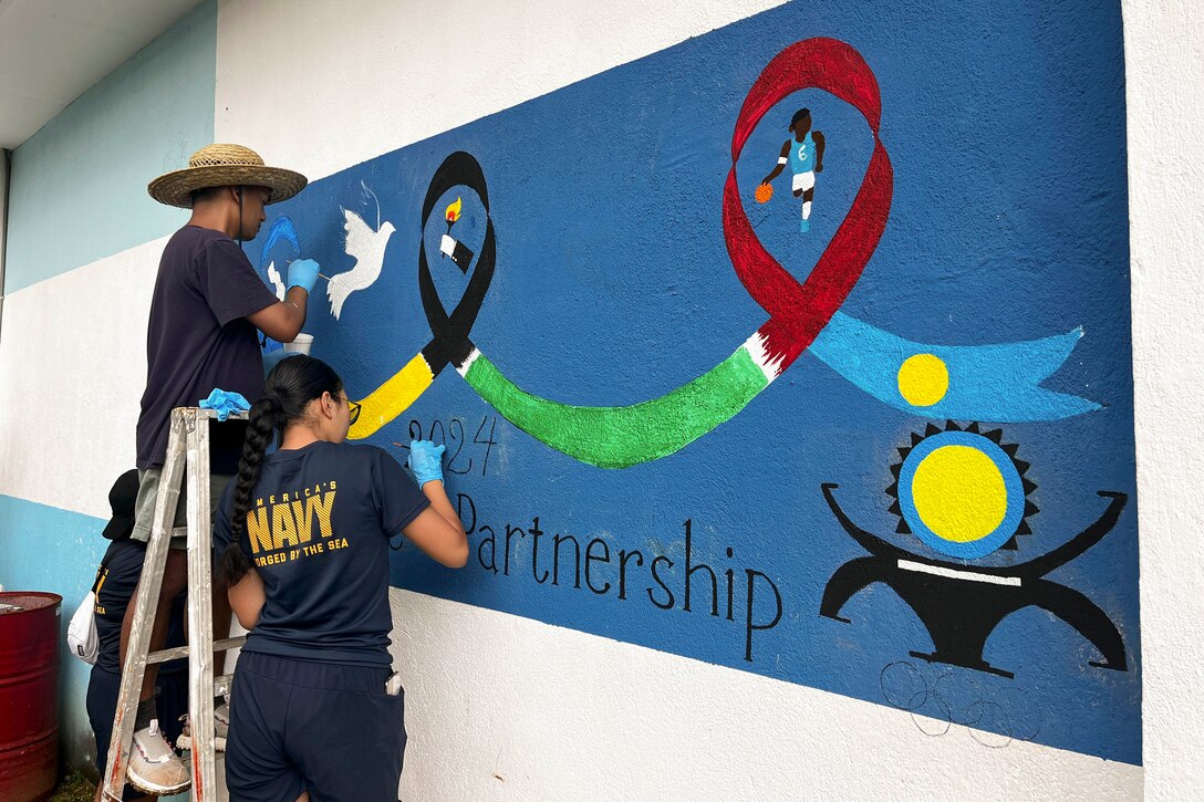 A sailor stands on a ladder as two others stand below while painting a mural on a building.