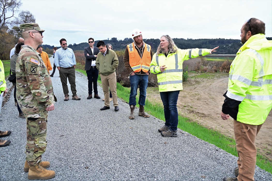 About 13 people stand on a paved road on top of a levee with farmland of behind them.