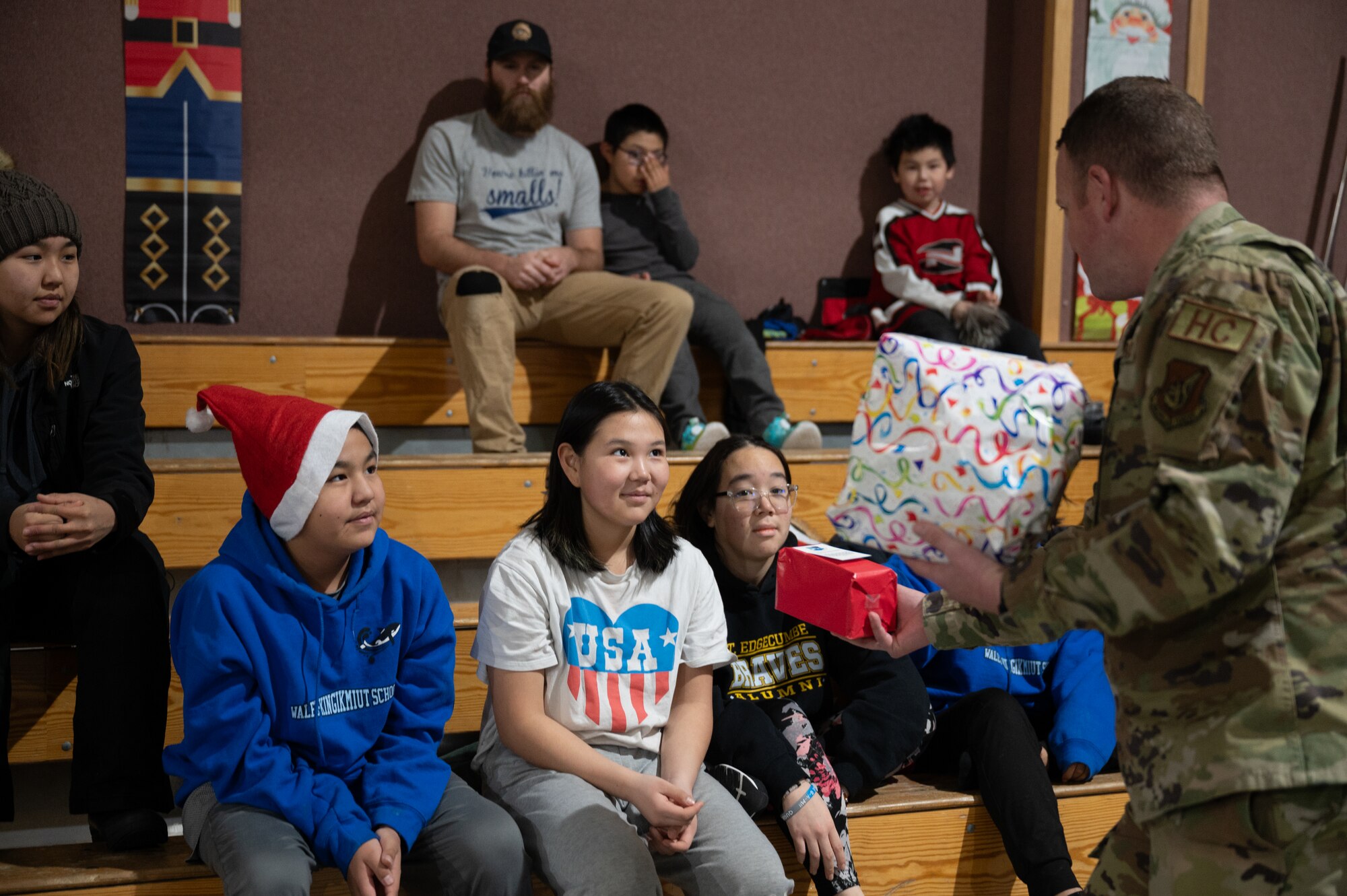 Parents, students, and teachers pose in the school gym with Pacific Air Forces Regional Support Center warriors for group picture, on Dec. 21, 2023, at Kingikmiut School, Wales, Alaska. The children each received Christmas gifts based on their age group. (U.S. Air Force photo by Airman 1st Class Quatasia Carter)