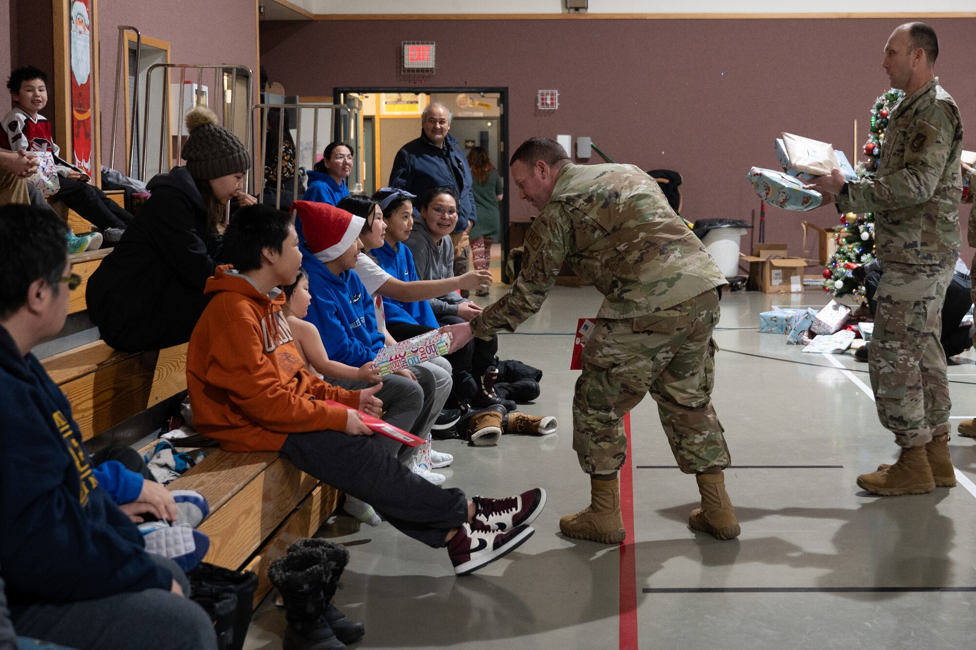 Parents, students, and teachers pose in the school gym with Pacific Air Forces Regional Support Center warriors for group picture, on Dec. 21, 2023, at Kingikmiut School, Wales, Alaska. The children each received Christmas gifts based on their age group. (U.S. Air Force photo by Airman 1st Class Quatasia Carter)