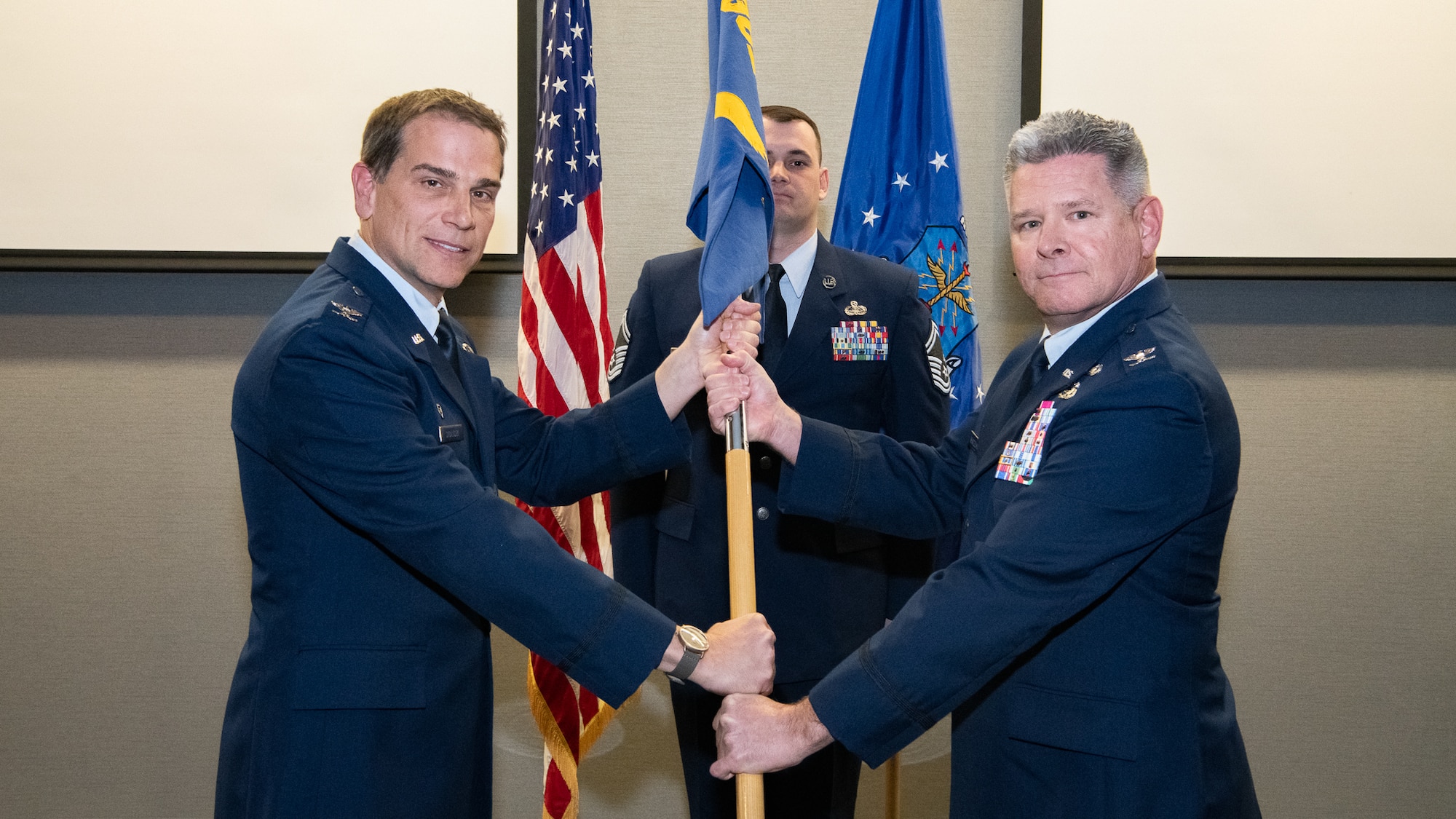 Two men in Air Force service dress grasp a flag pole while pausing for their photo to be taken.