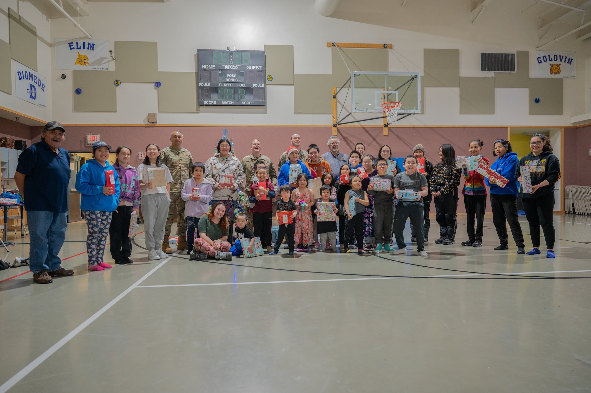 Parents, students, and teachers pose in the school gym with Pacific Air Forces Regional Support Center warriors for group picture, on Dec. 21, 2023, at Kingikmiut School, Wales, Alaska. The children each received Christmas gifts based on their age group. (U.S. Air Force photo by Airman 1st Class Quatasia Carter)