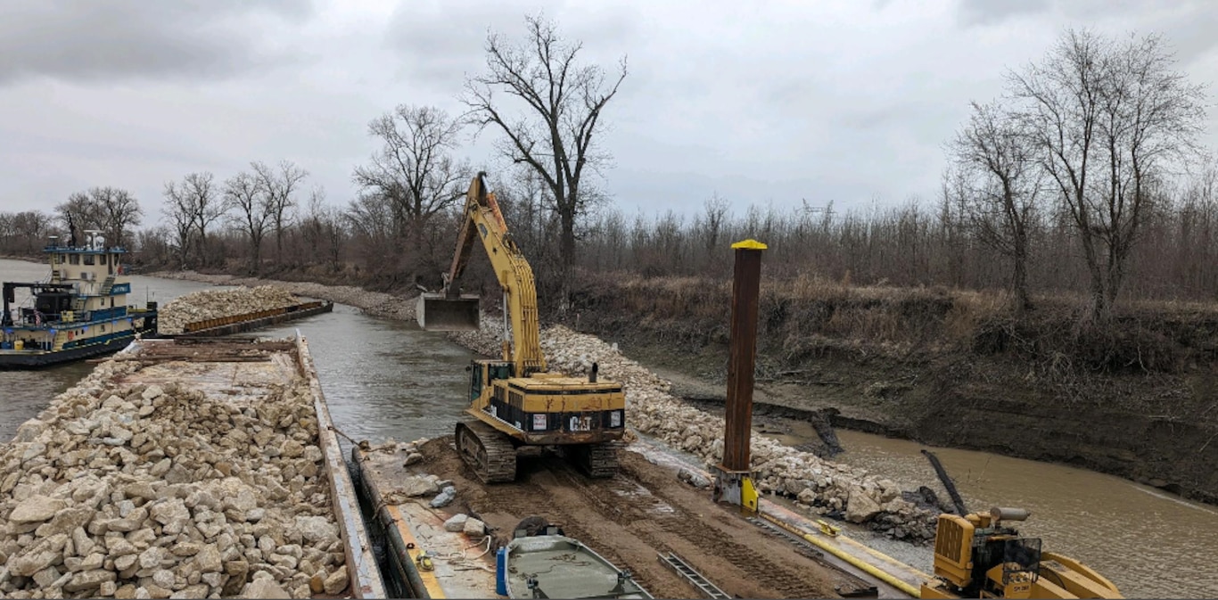 A construction boat places rock on the river in the bottom left with the river and trees in the background.