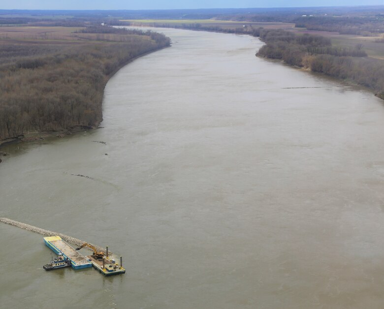 A construction boat places rock on the river in the bottom left with the river and trees in the background.