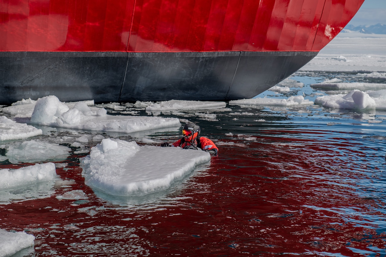 A Coast Guard member swims in icy water during rescue training. The hull of a Coast Guard cutter can be seen in the background.
