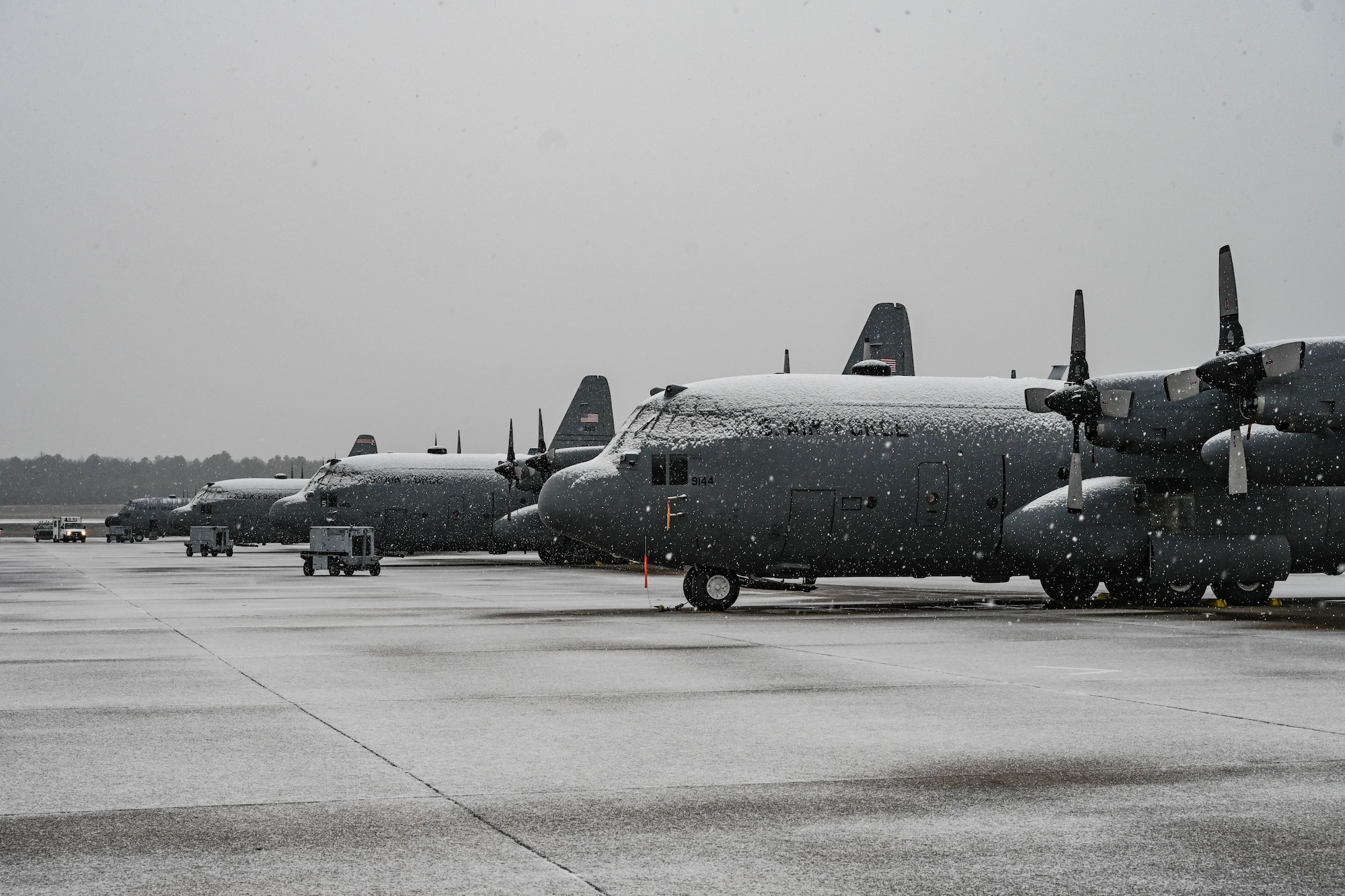A row of military aircraft covered in snow sit on a flightline.