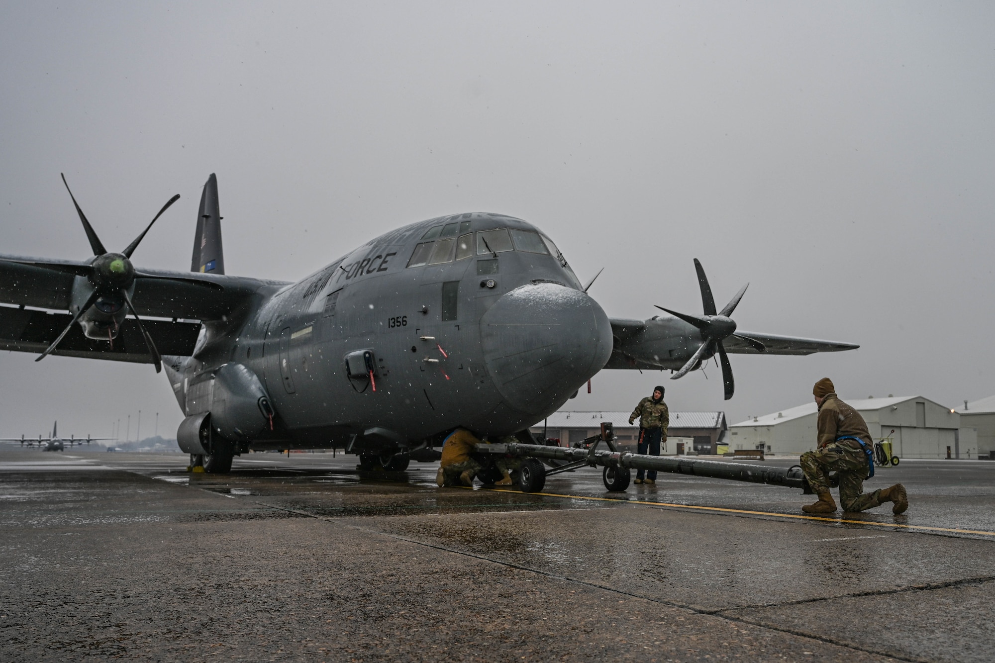 A military aircraft gets blanketed in snow while Airmen maintain it.