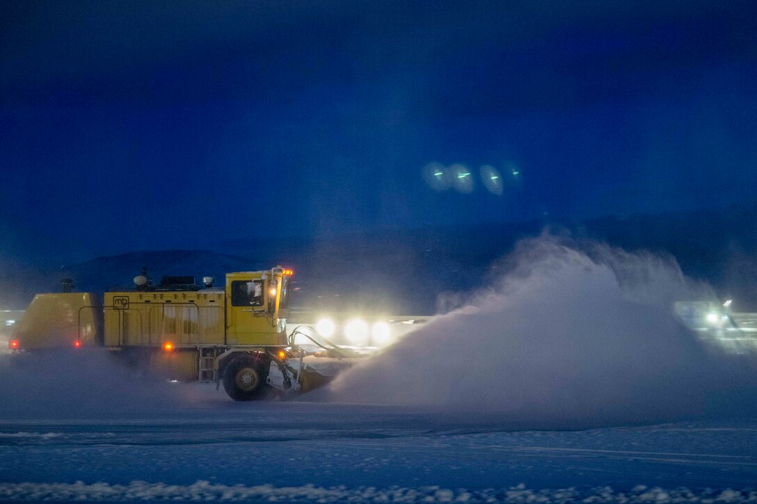 Snow flies in the air as Air Force personnel drive a snowplow truck to clear a path in the dark illuminated by headlights.