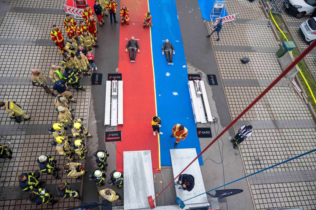 Dummies lay on the ground leading up to ropes hanging from the ceiling as dozens of firefighter gather on the side and watch as seen from above.