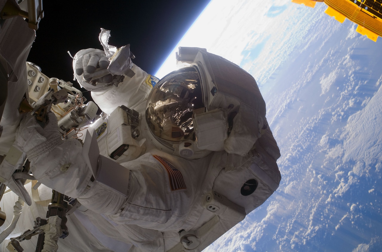 An astronaut works on a piece of the International Space Station while floating in space. Earth is seen in the background.