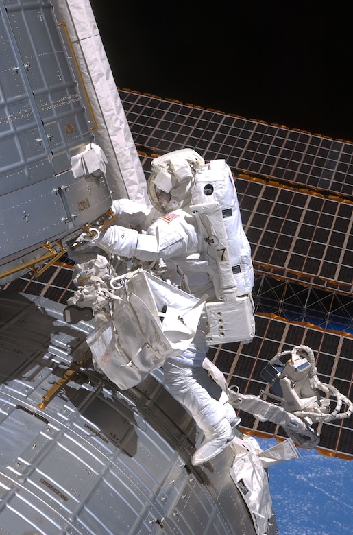 A person in a spacesuit, floats in space while holding onto a piece of a large space station.