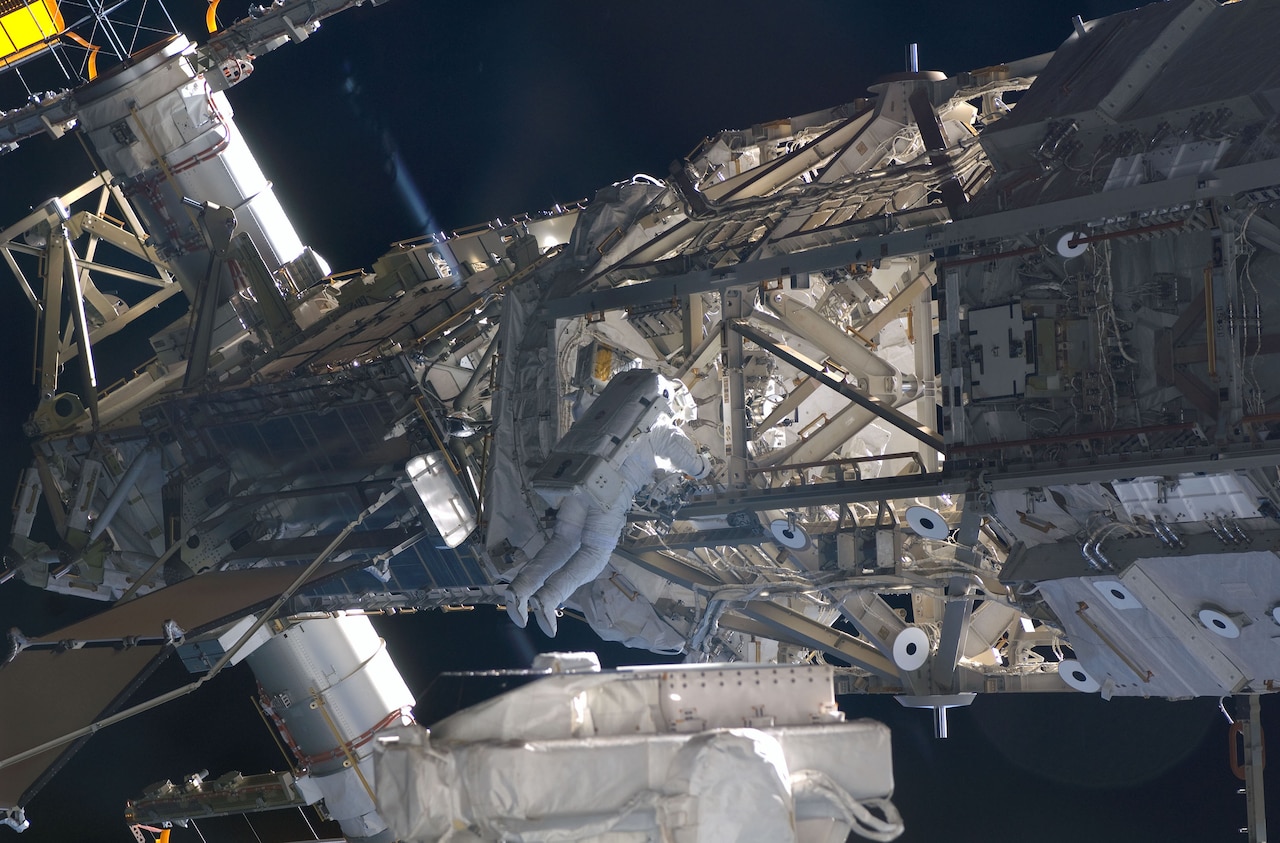 A person in a spacesuit hangs onto a part of a space station while in space.