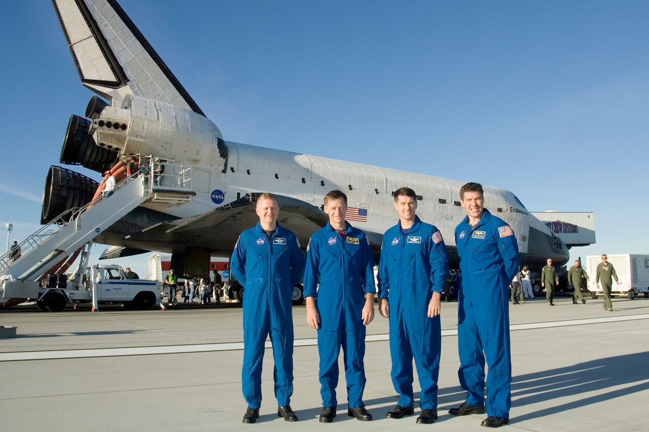 Four people stand on tarmac in front of a large space shuttle.