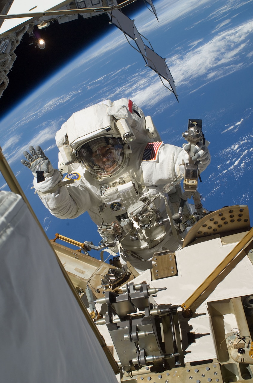 A person in a spacesuit, waves to the camera from the outside of a space station.  Earth is in the background.