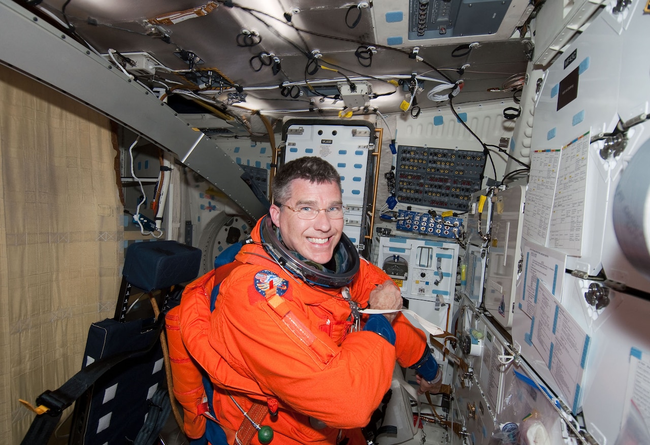 A person in a spacesuit smiles inside a small room filled with gadgets.