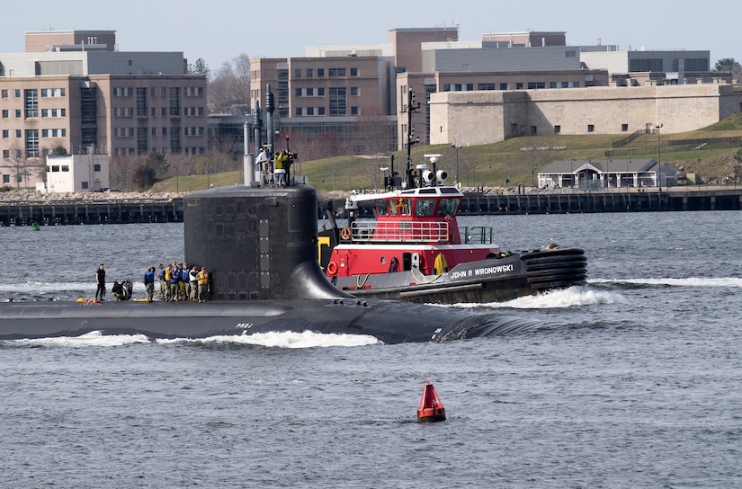 Sailors stand on the topside of a submarine as a towing vessel floats nearby.