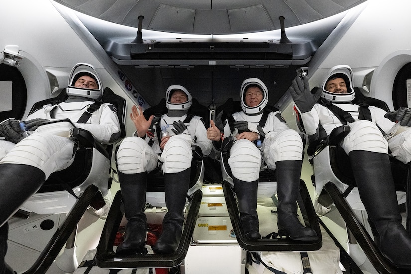 Four people in flight suits and helmets are strapped into seats.