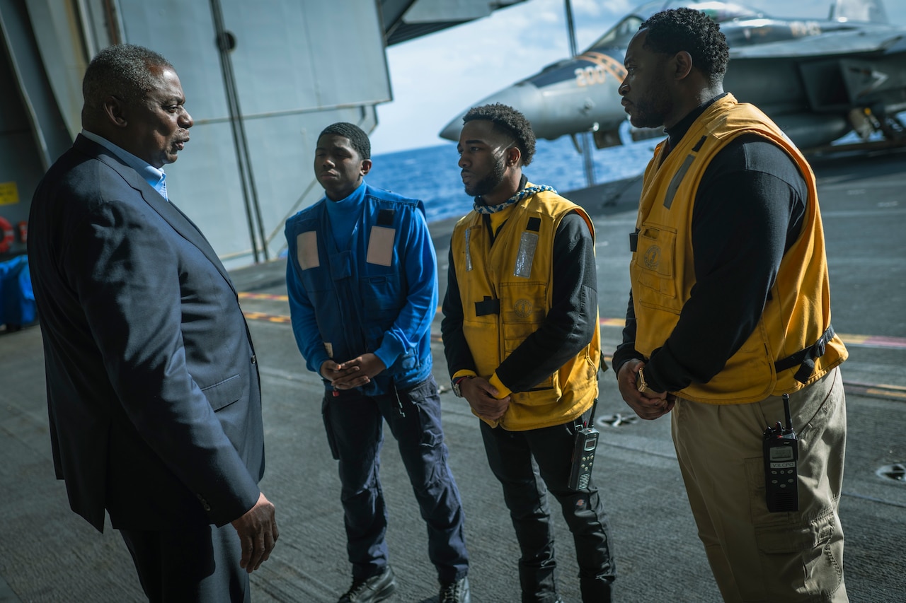 A man in a suit speaks with younger men who wear vests of varying color.  In the rear is a fighter aircraft.