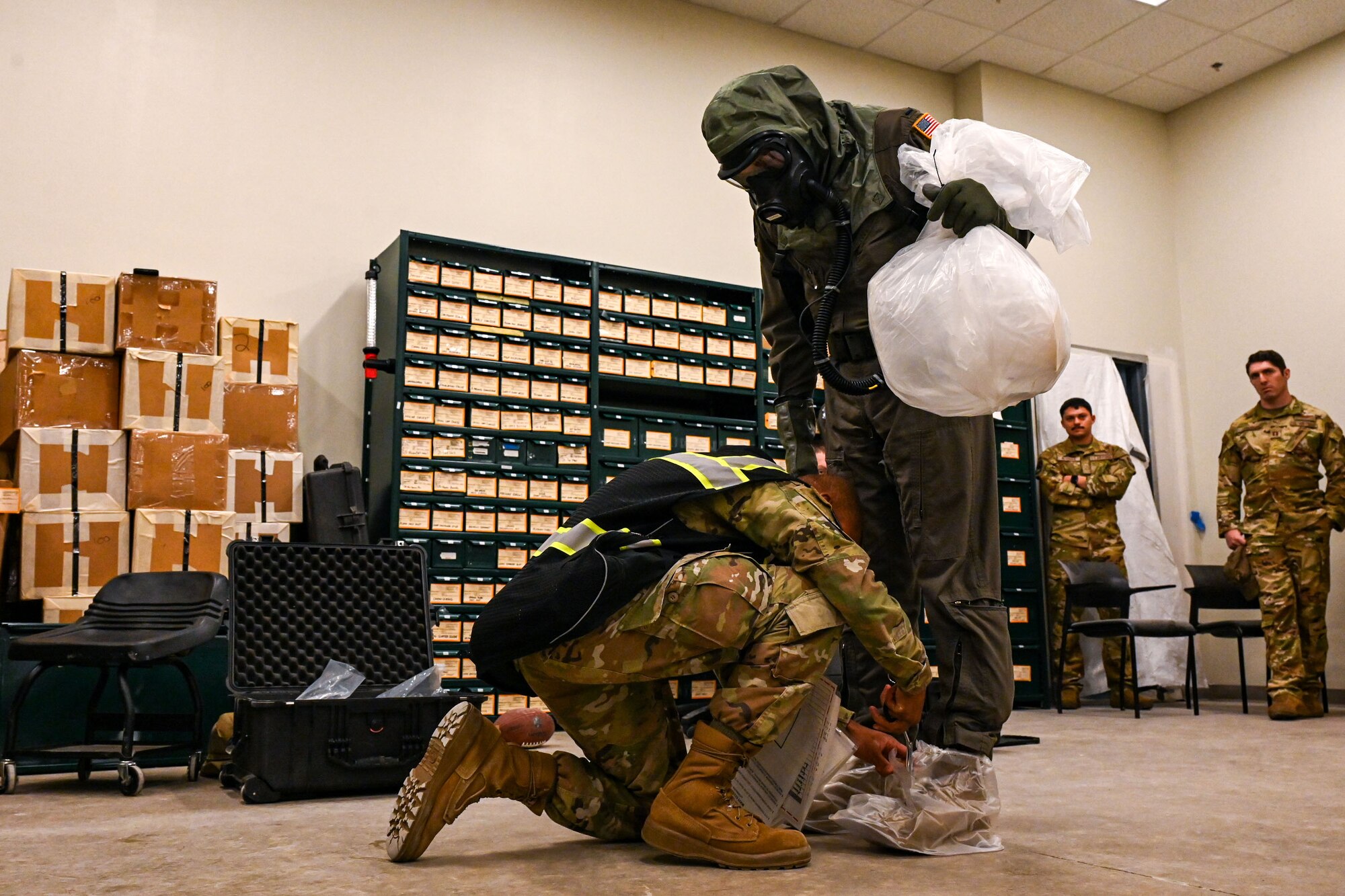 An Airman is kneeling and checking the fit of chemical safety gear around the ankles of a standing Airman.