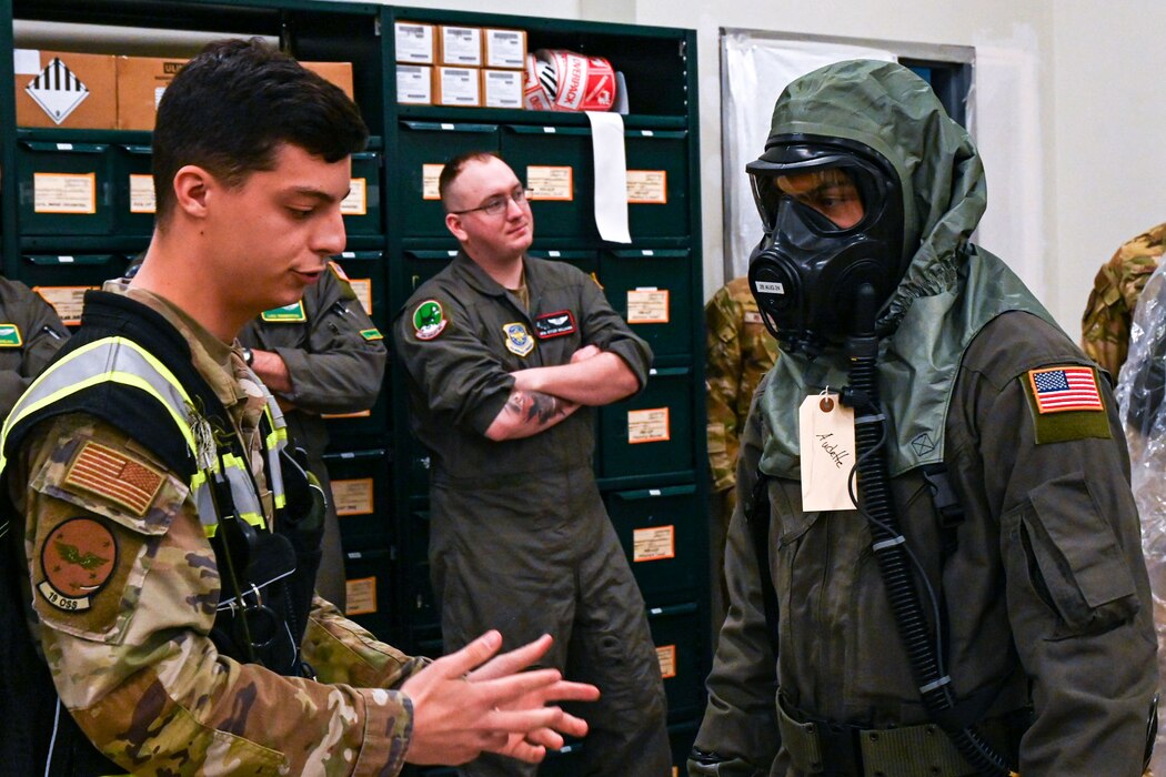 Airman speaks and makes hand gestures to another Airman in chemical safety gear.