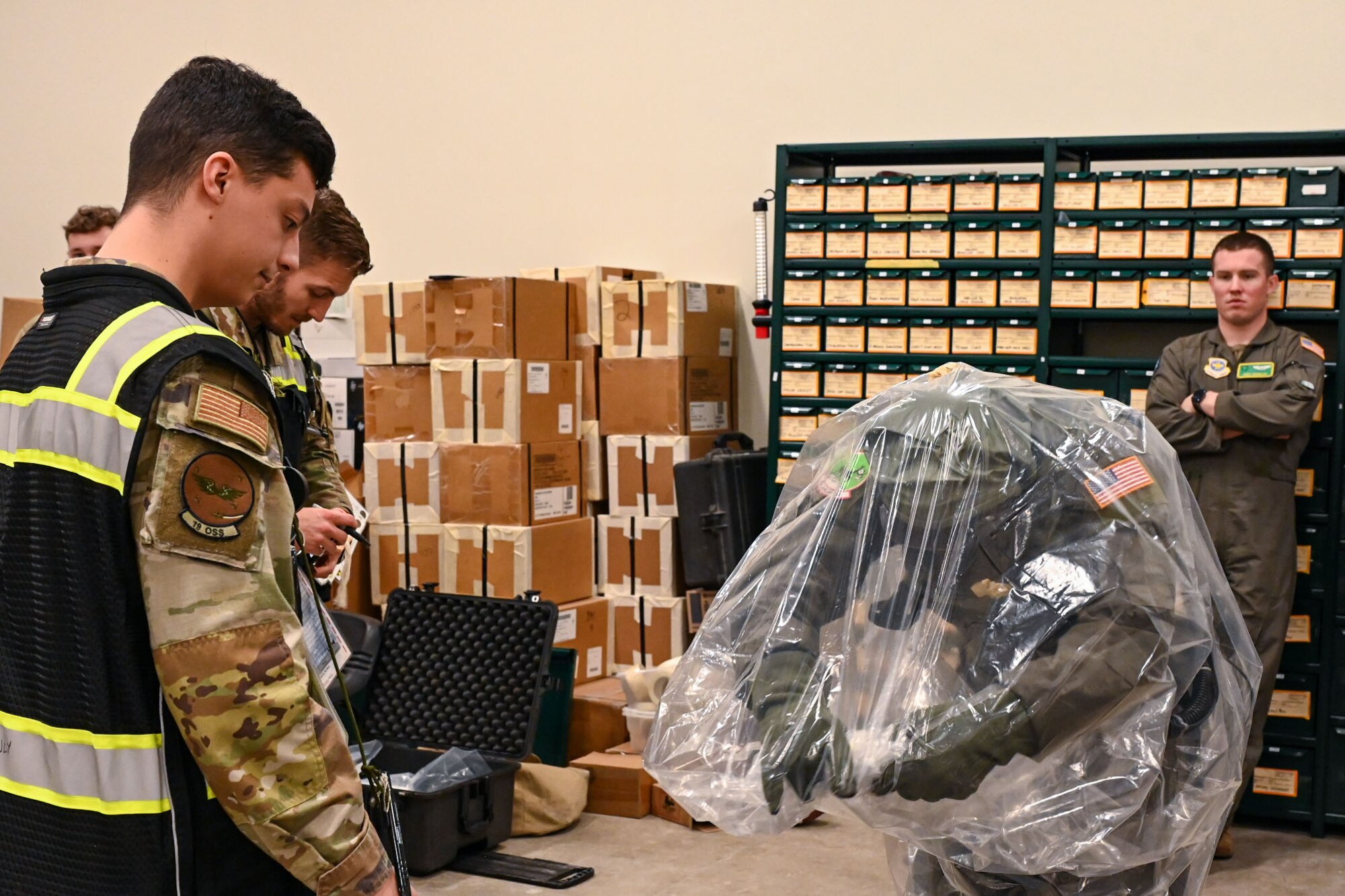 Airmen stand in a room and look at an Airman in contamination control gear.