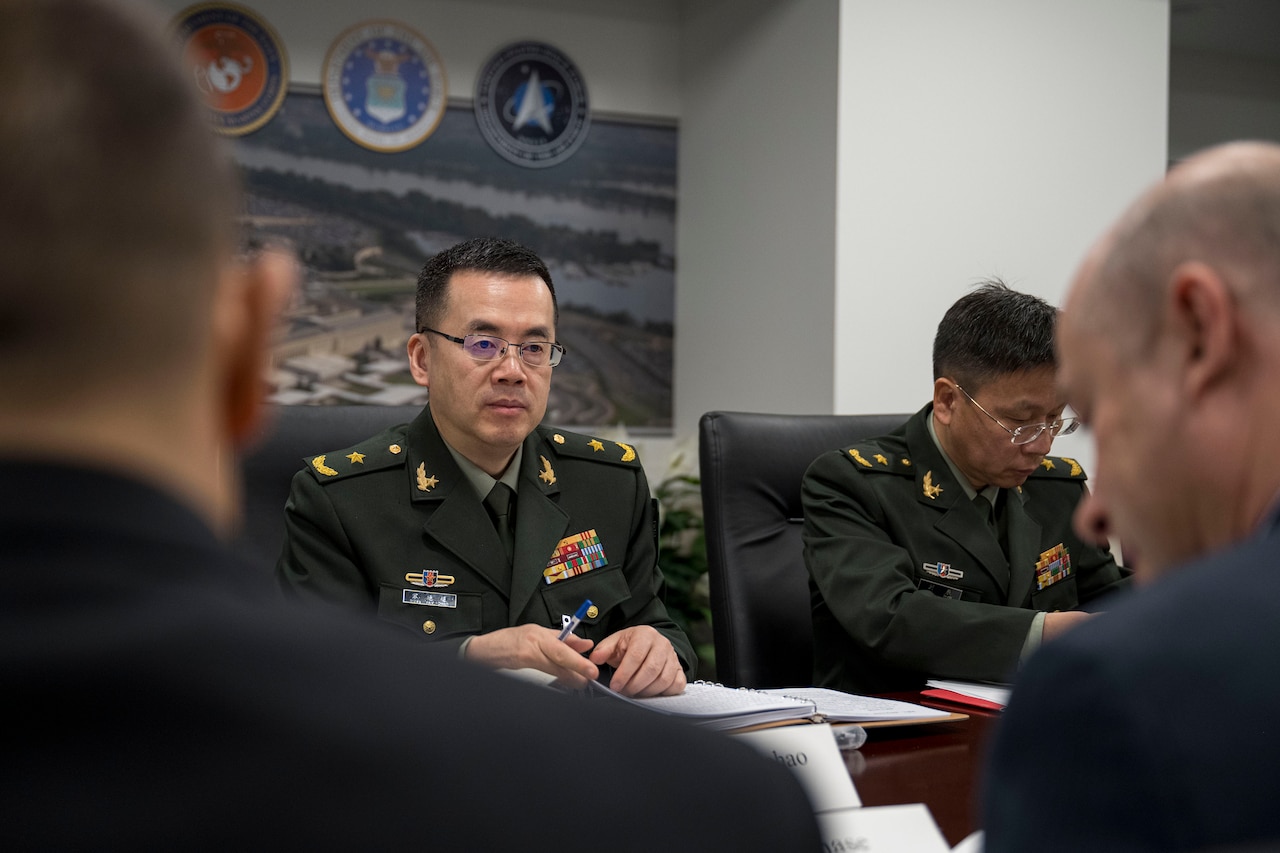 A person in a foreign military uniform is seated at a table across from a another person in a business suit.