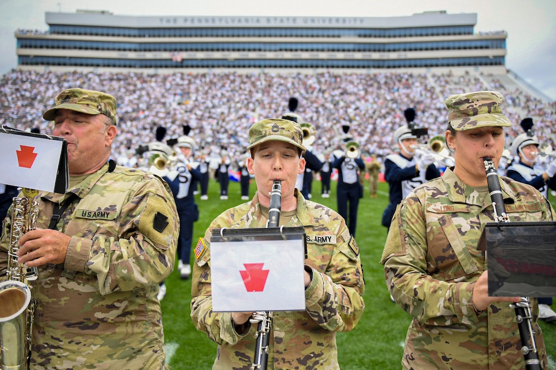 A close-up of three soldiers playing instruments with a college marching band performing in front of packed stadium in the background.