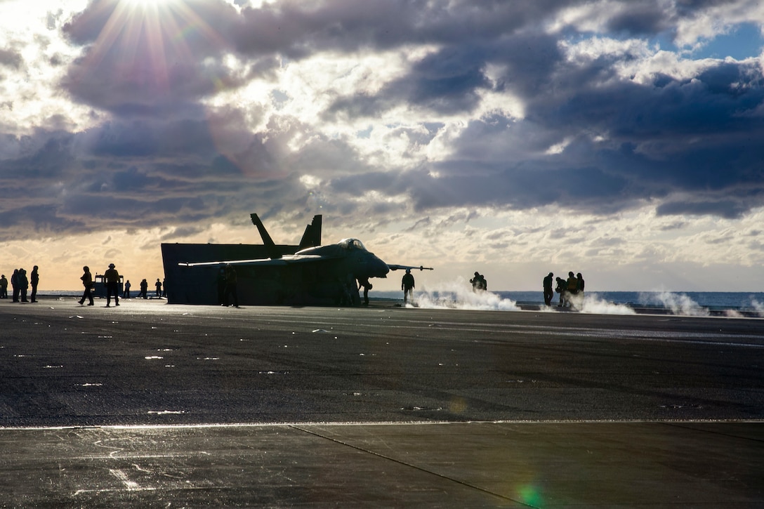 An aircraft surrounded by sailors prepares to take off aboard a ship at sea under a sunlit sky.