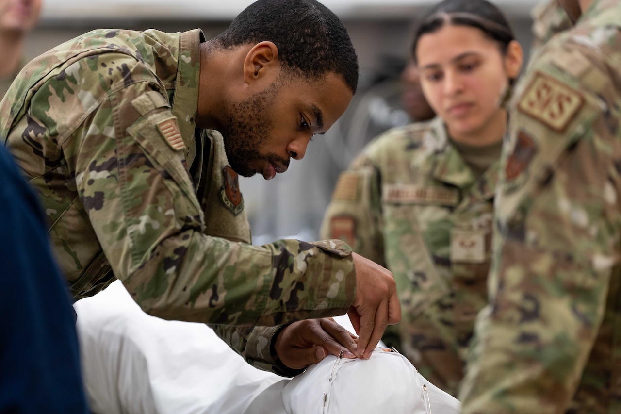 An airman  pins the folds of a white sheet covering a training mannequin while another airman observes.