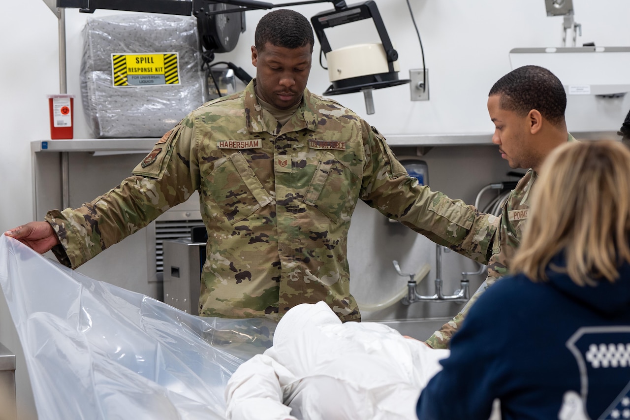 An airman holds up the edges of plastic wrapper with a training mannequin on top while two others look on from the side.