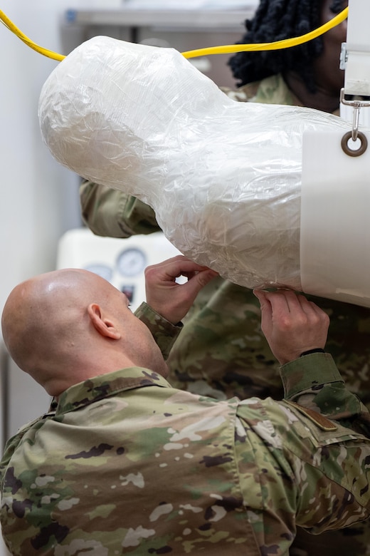 An airman tucks the edges of plastic wrapping beneath the shoulder of a training mannequin from below.