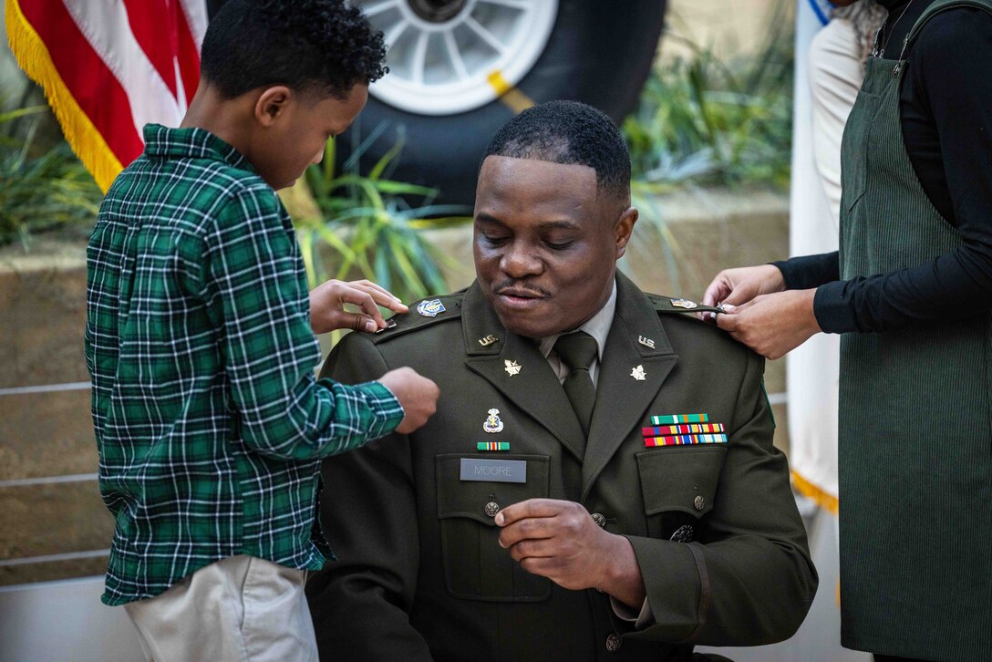 A child and another family member pin insignia on the shoulders of an Army officer.