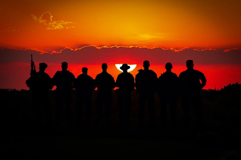 A silhouettes photo illustration of eight soldiers standing in a row with a vivid red and orange sky behind them.