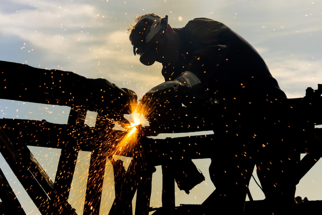 A Marine in protective head gear cuts metal, causing sparks to fly.