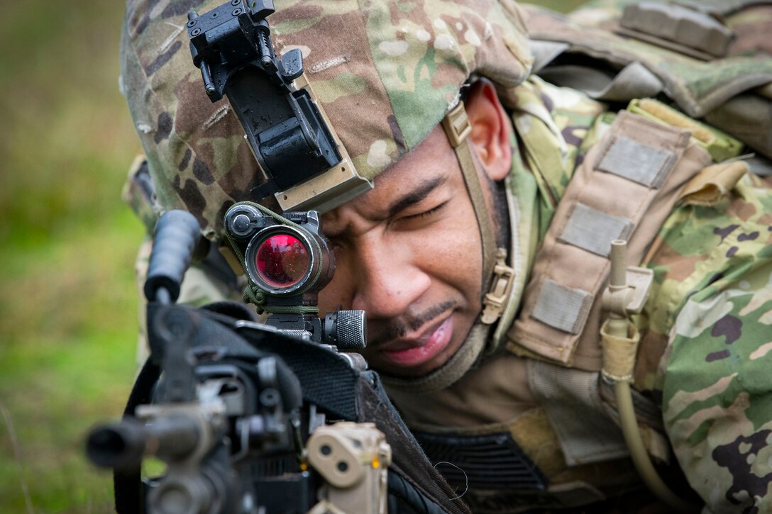 A guardsman wearing tactical gear lays in the grass while looking through a gun’s scope.